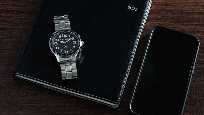 Picture of a black notebook and a smart phone. On top of the notebook is a GNSS voice watch.